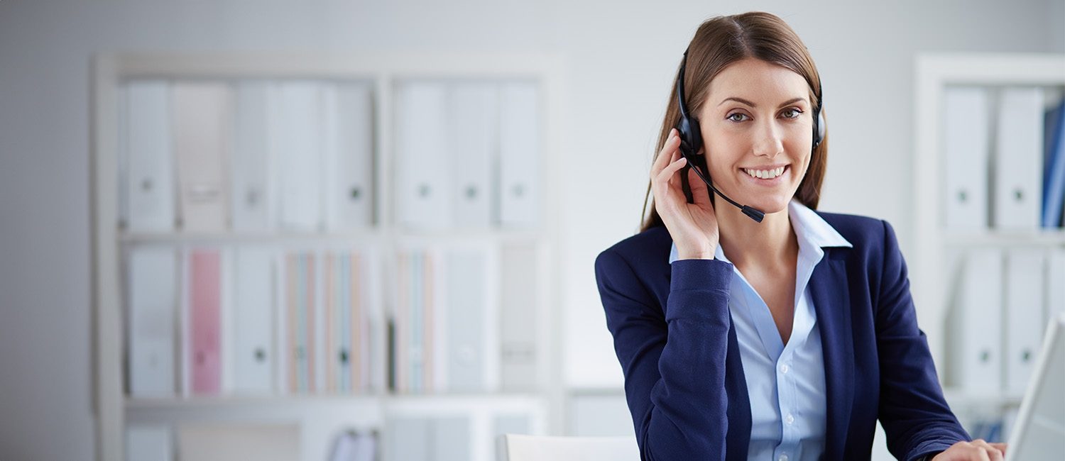 Una operadora de call center sonríe amablemente mientras mira su monitor, con una mano teclea y con la otra sostiene el mouse de su computadora, el fondo está fuera de foco​