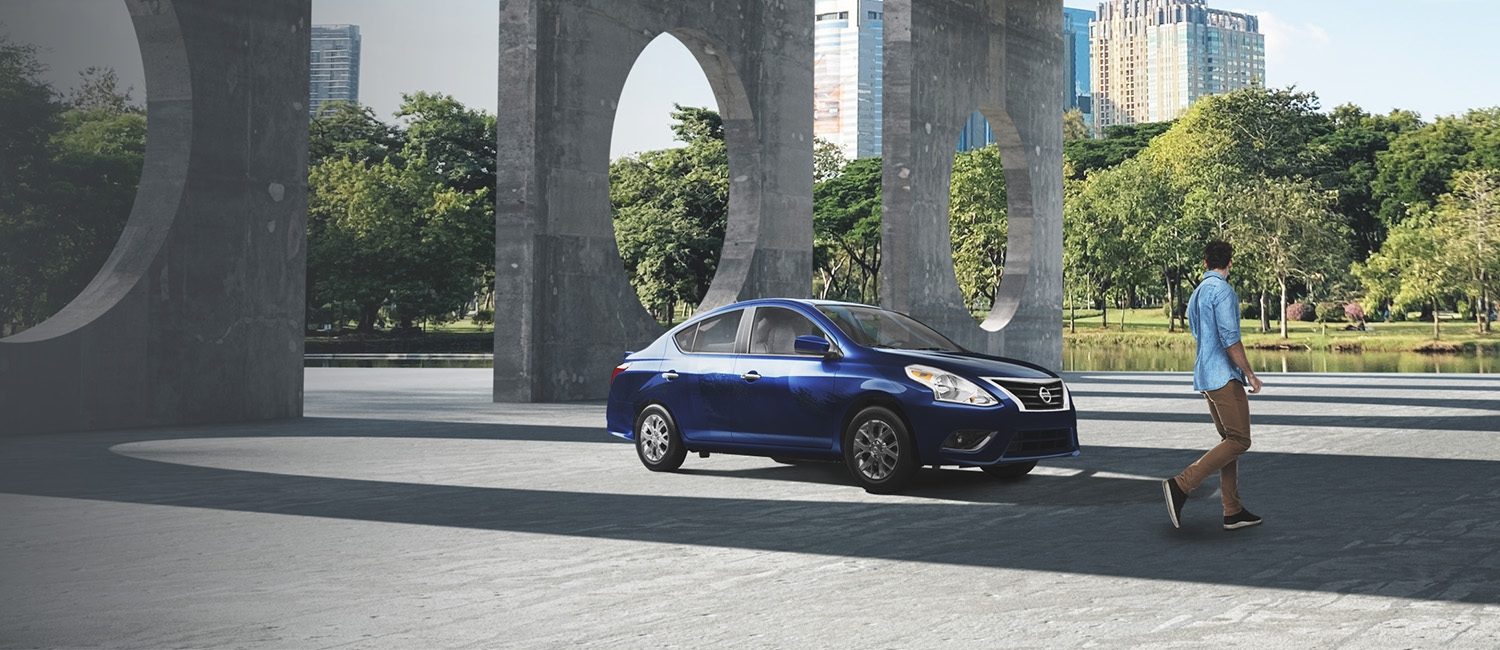 Un hombre camina frente a un Nissan Versa de generación anterior mientras lo mira en un parque citadino